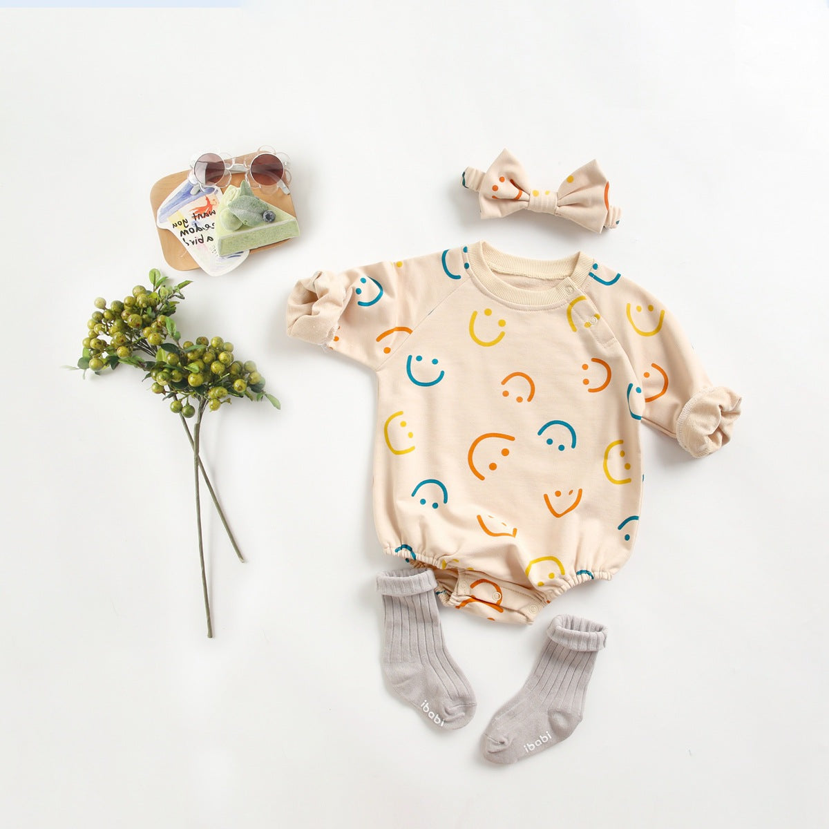 Beige baby romper met kleurrijke smiley-print, gecombineerd met bijpassende strik, sokjes en accessoires, flatlay gefotografeerd op een witte achtergrond.