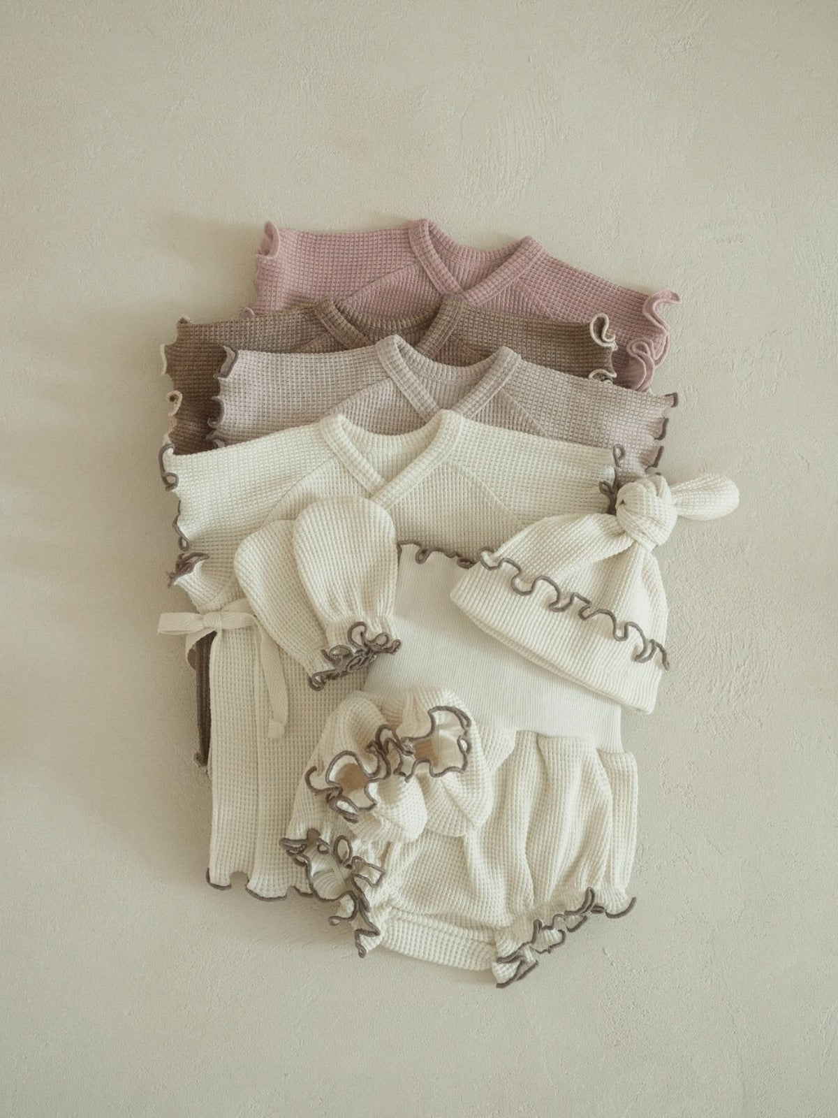 Gestapeld unisex babyzomersetje in waffle-stof met monk-style top, bloomer en muts in beige, taupe en roze tinten, gefotografeerd tegen een zachte crèmekleurige achtergrond.